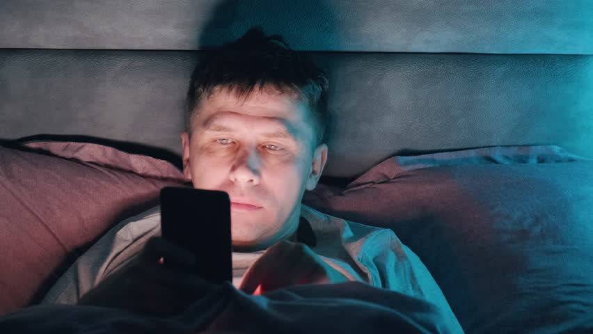Man with mobile phone lying on his bed in the dark while scrolling smartphone with illuminated face. Surfing internet, social media at night. Insomnia concept