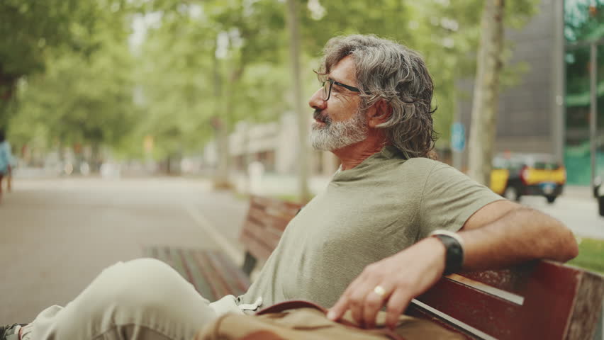 Positive middle-aged man with gray hair and beard wearing casual clothes sits on bench. Mature gentleman in eyeglasses is resting on bench crossing his fingers