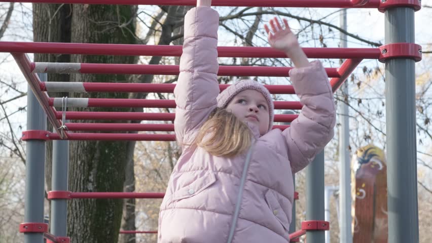 School girl climbs on horizontal bars on the playground in spring park. A sporty child does exercises on monkey bars in kindergarten. Children sports and activities. Healthy lifestyle | Shutterstock HD Video #1101121197