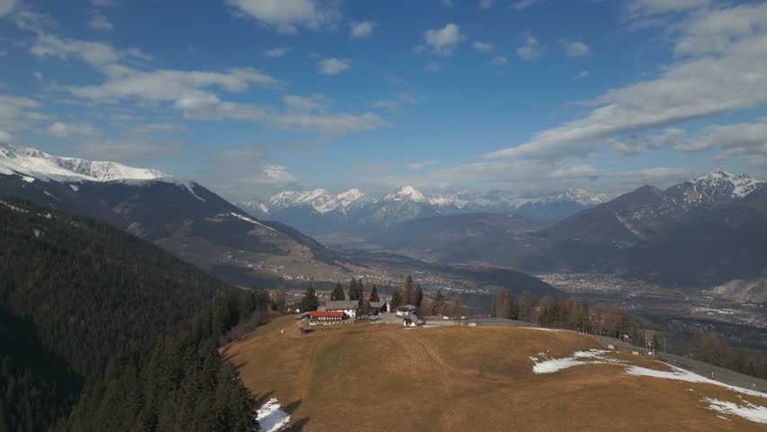 Aerial drone flying over houses on a mountain with fields and trees besides. A big valley with alpine mountain ranges and a city in the distance. Flying forward on a sunny winter day. Tyrol, Austria