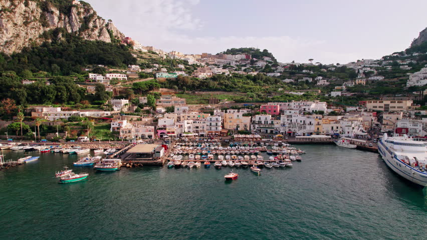 An ascending aerial shot from the boats in busy Marina Grande, over stunning cliffs and winding roads up to the boutique shops, fashionable cafés and luxury hotels of Capri Town in Campania, Italy.