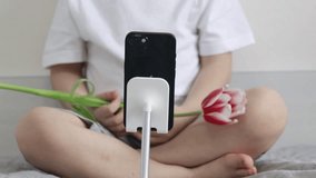 young blogger vlogger kid child sitting with smartphone on holder in front talking with flowers holding one tulip in hands gesturing.modern pre schooler boy close up feet using modern device