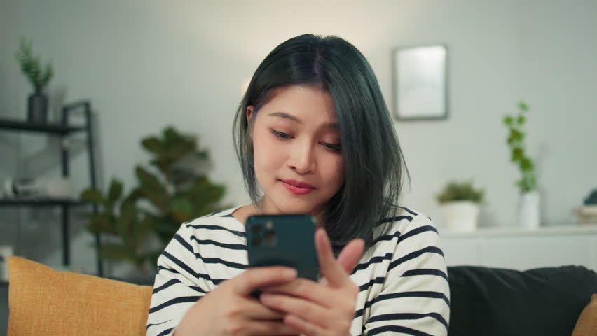 Young Asian woman holding mobile phone in hand playing social media. Beautiful girl looking at smartphone surfing internet, chatting with friends. Happy smiling female using cellphone in living room. | Shutterstock HD Video #1101126979