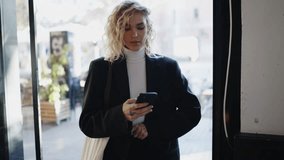 Attractive blonde hair female standing with mobile phone device while reading text message. Concentrated young woman typing email on smartphone application. Slow motion