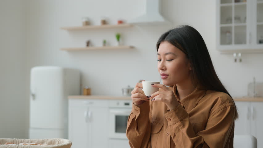 Relaxed girl Asian korean woman in domestic kitchen with cup of coffee drinking hot herbal tea multiethnic lady thoughtful calm housewife drink beverage looking in distance dreaming with mug in hands Royalty-Free Stock Footage #1101131087