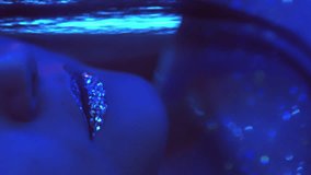 Vertical video. Disco makeup. Neon girl. Nightclub beauty. Cropped closeup of blue color light woman face with sparkling glitter skin metallic lips on dark.