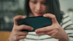 Close Up of Serious Young Beautiful Asian Woman Hand Holding Smartphone Smile Focus on Playing MOBA MMORPG Mobile Game Enjoy Looking and Tapping At Phone Screen Seated on Couch in Living Room.