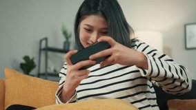 Happy Attractive Asian Woman Hand Holding Smartphone Smile Focus on Playing MOBA MMORPG Mobile Game Looking at Cellphone screen Express Joyful Excited and Cheerful Face sit on couch in Living Room