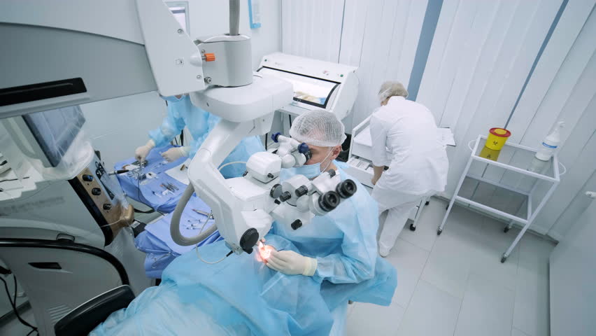 Ophthalmological surgery specialist sits in front of microscope performing operation. Low angle view on the female doctor conducting high-precision surgery. Femto Laser Assisted Cataract Surgery. Royalty-Free Stock Footage #1101138483