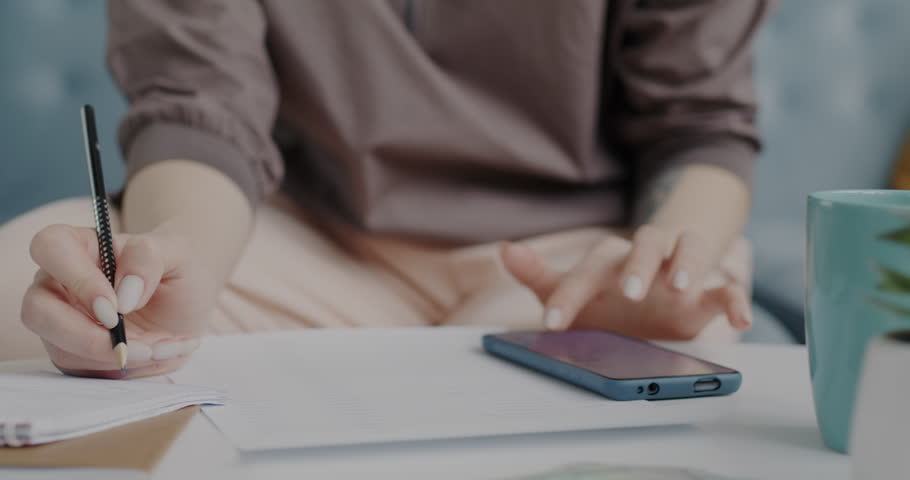 Close-up of female hands using smartphone and writing notes on paper indoors at home. Modern technology and business paperwork concept.
