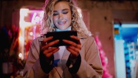 Female with white teeth smile stands on night city street with neon lights  while reading message on mobile phone. Millennial young woman using smartphone app and watching content in social media
