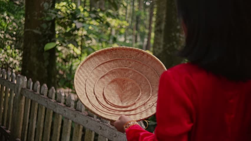 Over-the-shoulder shot of an Asian woman in a red traditional dress, tossing a conical straw hat onto the floor of a wooden bridge in a green forest.