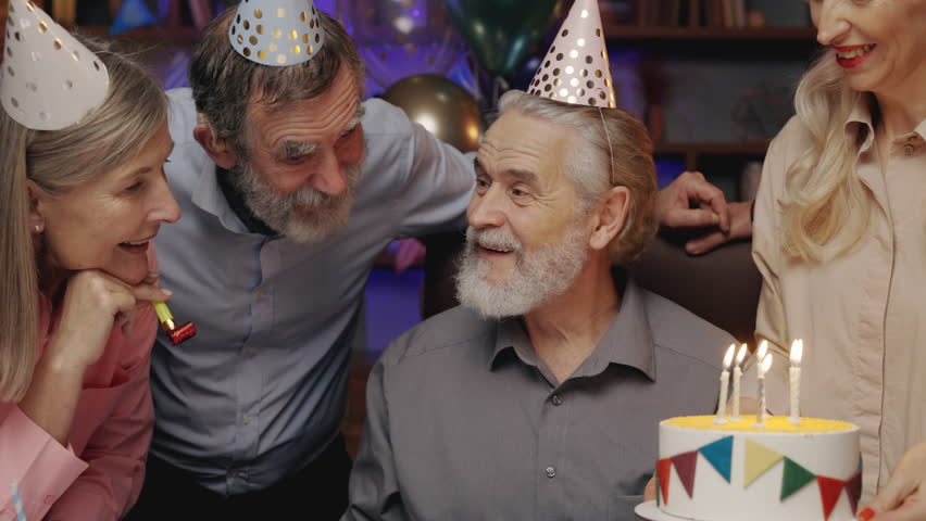Elderly Man in Birthday Hat Blowing Out Candle on Birthday Cake, Celebrating Together with Other Senior People at Nursing Home. Birthday Party, Old Friends Blowing Party Horns. S3niorLife Royalty-Free Stock Footage #1101142967