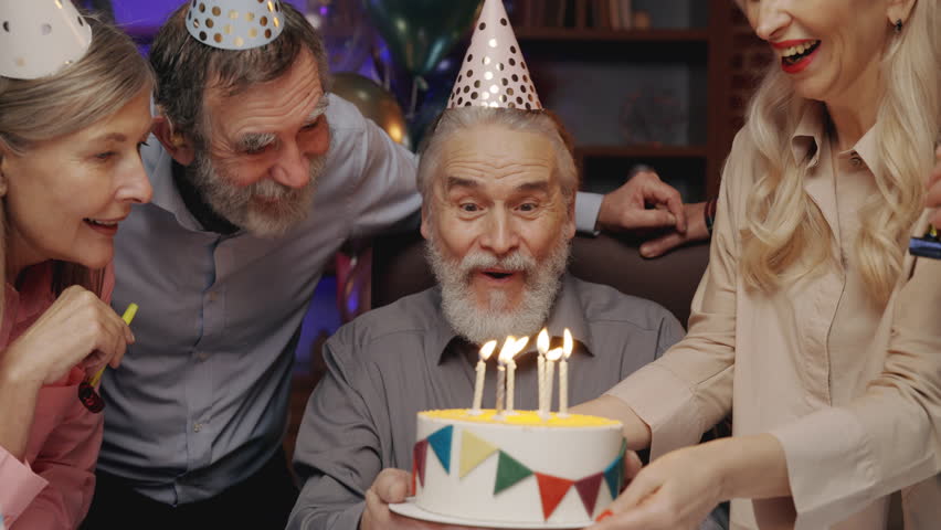 Elderly Man in Birthday Hat Blowing Out Candle on Birthday Cake, Celebrating Together with Other Senior People at Nursing Home. Birthday Party, Old Friends Blowing Party Horns. S3niorLife