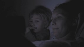 Mom and son lie in bed with the lights off and watching cartoons from the phone