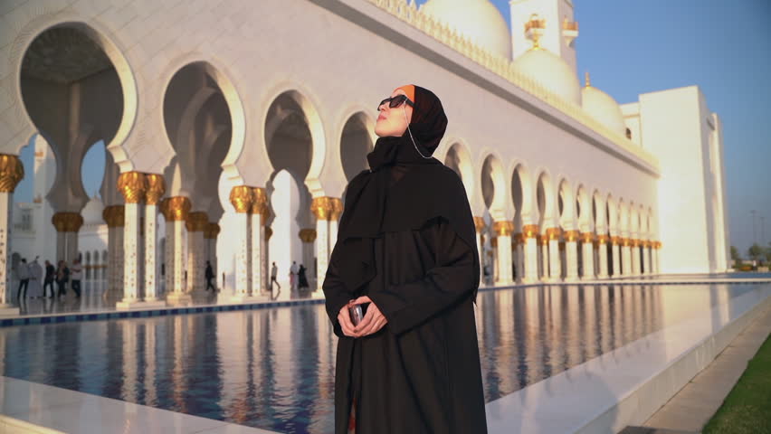 Woman in hijab examines mosque in UAE while traveling spbas. Closeup view of beautiful young female looking at white architecure and posing with smile, holding phone and standing outdoors. Attractive