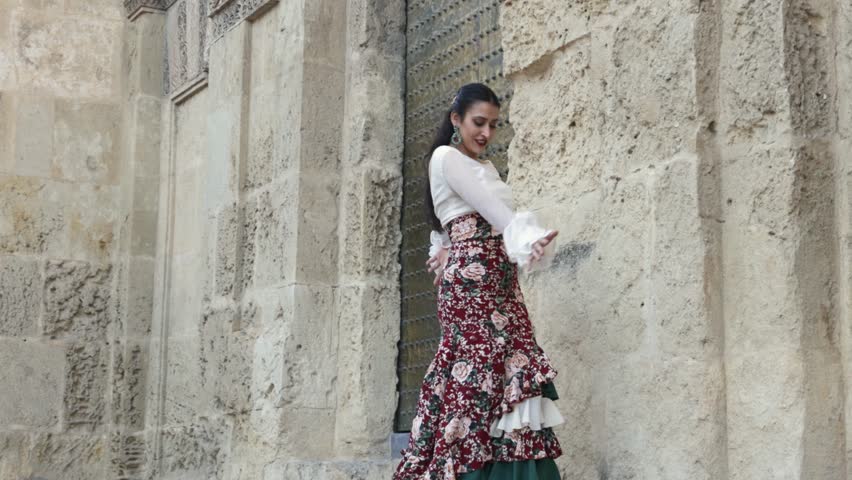 Hispanic gypsy woman flamenco dancer dressed in typical flamenco dress with red flowered skirt and white shirt dancing next to a wall of an old building Royalty-Free Stock Footage #1101153623