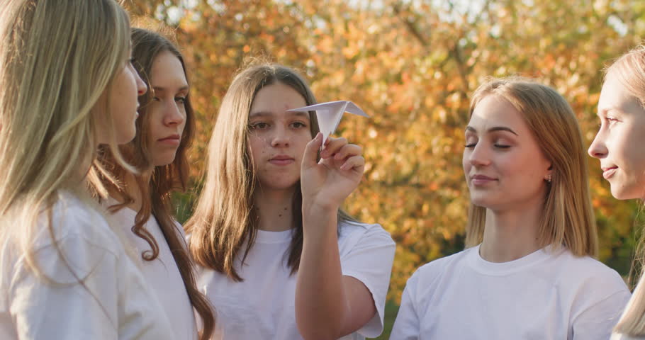 Group of teenagers girls holds single paper airplane preparing to launch in city park illuminated by sunlight. Teenage schoolgirls hold airplane smiling