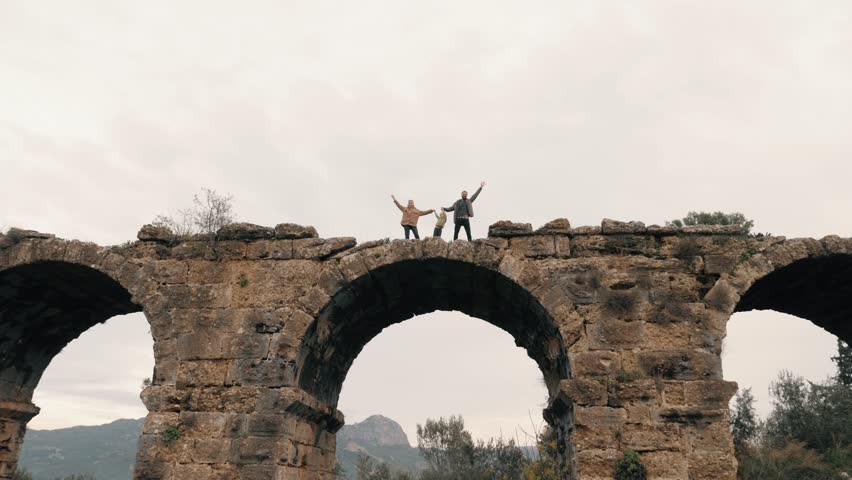 Happy travel family posing on top of antique arch ruins construction greeting waving hands tourist mother father and son enjoy journey adventure destination explore sightseeing spending time together