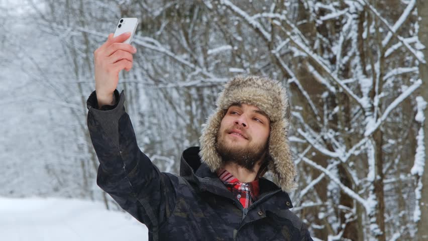 Young Caucasian male blogger in fluffy hat with earflaps stands in winter forest and takes selfie video or photo on phone. Friendly charismatic guy makes various grimaces on his face.