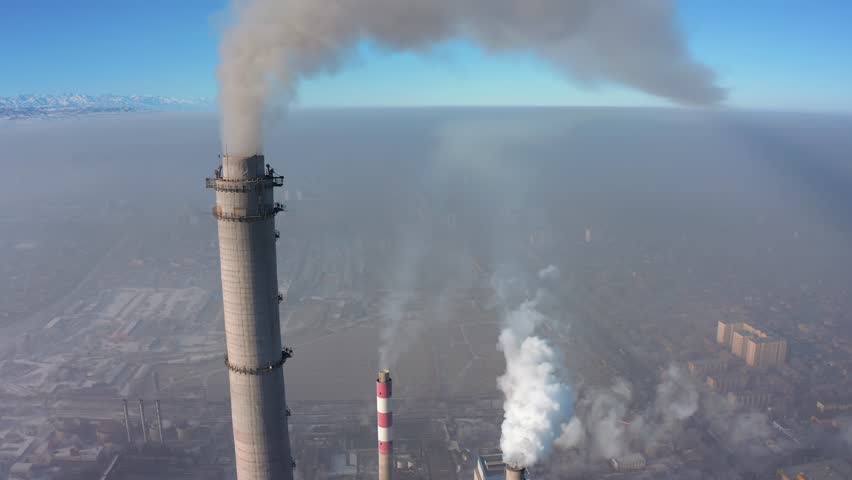 View of the smoking top of a long chimney of a factory polluting the environment against the backdrop of a city covered in gray smog and a blue sky