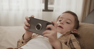 Kid using phone for gaming. Child playing video game indoors. Beautiful child boy 6 years old playing mobile game on smartphone at home. Adorable kid boy playing mobile phone. 4K