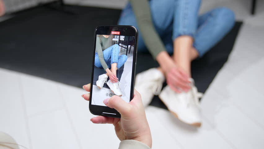 Taking a video of a model on a smartphone. Girl at the backdrop sits on the floor tying her sneakers.
