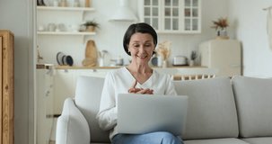 Cheerful excited senior woman talking on video call at home, sitting on couch, keeping laptop on lap, waving greeting hand hello at camera, chatting online, speaking, smiling, laughing