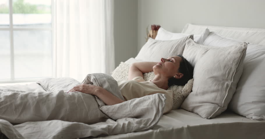 Carefree peaceful mature woman waking up on orthopedic mattress, resting in white bedding, sitting up, stretching body, rising hands, enjoying good morning after healthy sleeping Royalty-Free Stock Footage #1101170151
