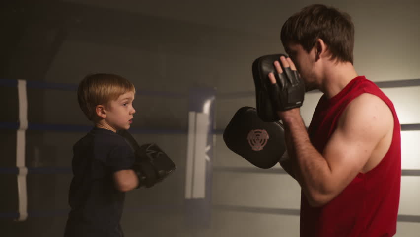 Portrait of a skilled boxer teaching little boy to fight in boxing gloves. Close-up view of adult man exercising in sparring with young kid in the ring with ropes. High quality 4k footage Royalty-Free Stock Footage #1101178671