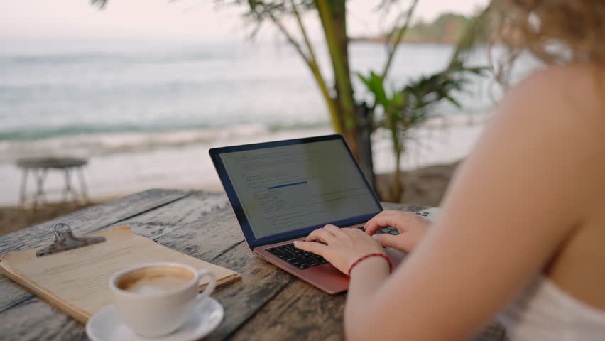 Female developer working on laptop by the ocean. Young woman freelancer coding at outdoor tropical cafe. Caucasian girl working remotely typing on computer at exotic location. Worldwide work concept. Royalty-Free Stock Footage #1101187855