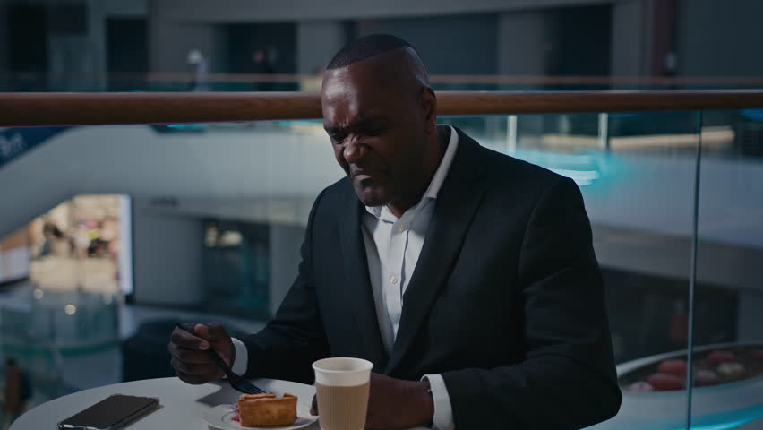 African American middle-aged ethic businessman entrepreneur man in cafe sitting at cafeteria table eating dessert dissatisfied with bad taste cake disgust pie drink nasty tea tasteless coffee drinking