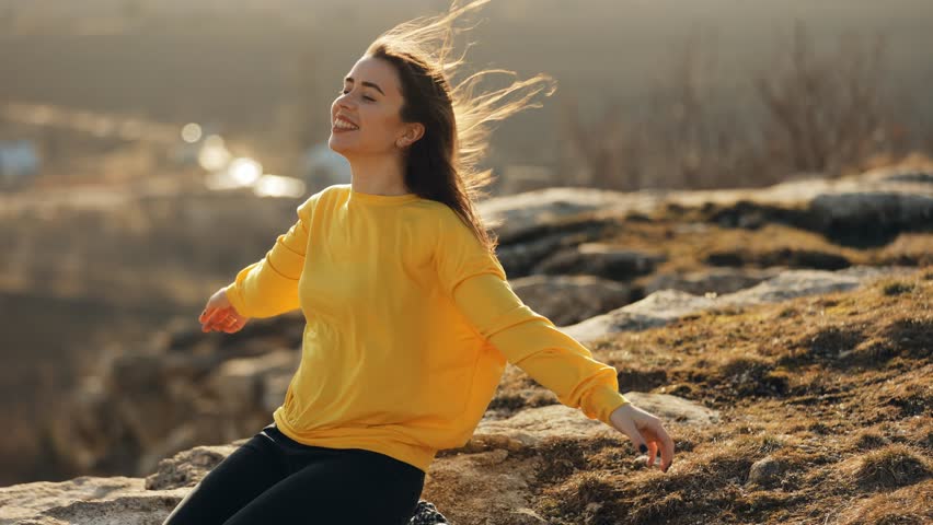 A young woman in a yellow sweater sits on the edge of a rock and raises her arms to the sides imitating flight, happy and cheerful admiring nature, wind blowing her hair in the sunlight, traveling alo