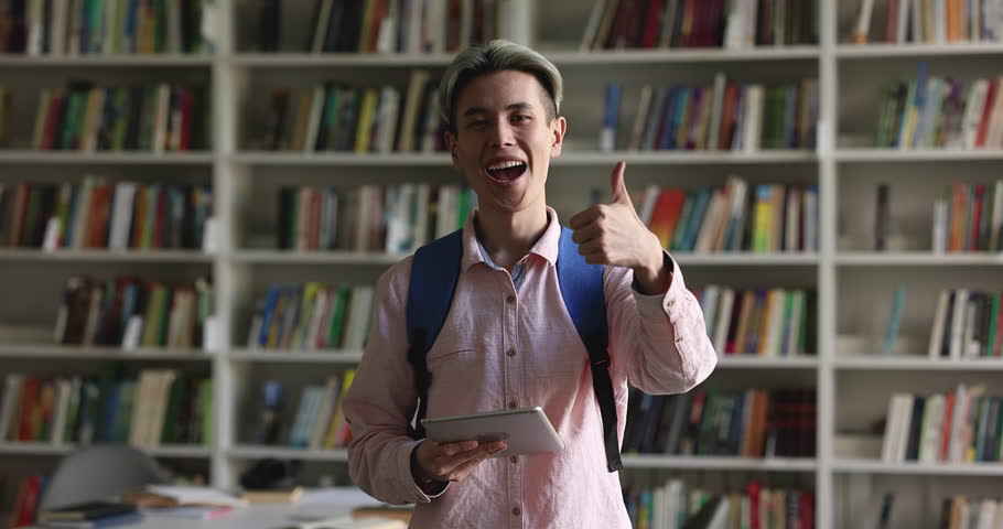 Happy Asian student guy holding tablet, reading on screen, standing in public school library, rising hand, showing like hand at camera, making thumb up gesture, smiling, laughing