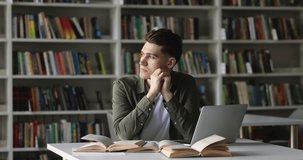 Serious college student guy thinking on project, looking away, using laptop in library, reading open books, textbook, writing notes, summary, working on research study, class report