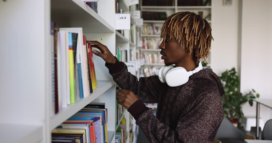 Serious African college student with wireless earphones searching literature on bookshelves in university library, picking book for research study writing, preparing for graduation exam. Side view Royalty-Free Stock Footage #1101193187