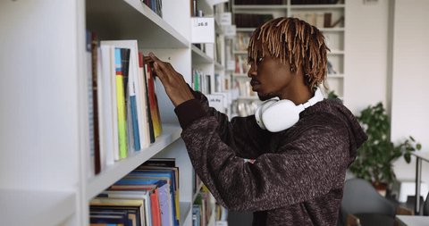 Serious African college student with wireless earphones searching literature on bookshelves in university library, picking book for research study writing, preparing for graduation exam. Side view - Βίντεο στοκ