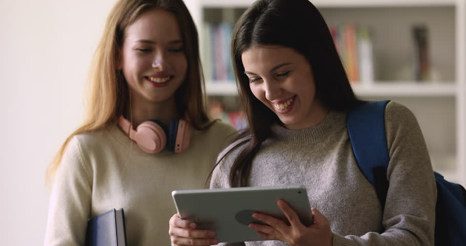 Two happy classmates girls talking in library, standing together, smiling, laughing, watching online content on digital gadget. Cheerful student girl showing presentation on tablet to friend
