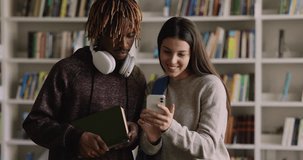 Multiethnic couple of cheerful students using mobile phone in library together, browsing Internet. Latin college, high school girl showing content on cellphone to African classmate guy in library