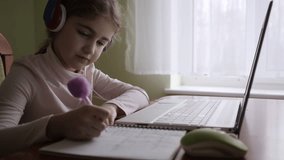 School Student Wearing Headphones Watching Internet Video Course. Distance Learning. Schoolgirl Studying Online Using Laptop Making Notes in Copybook. Caucasian Girl Wear Headphones Learn Online Class