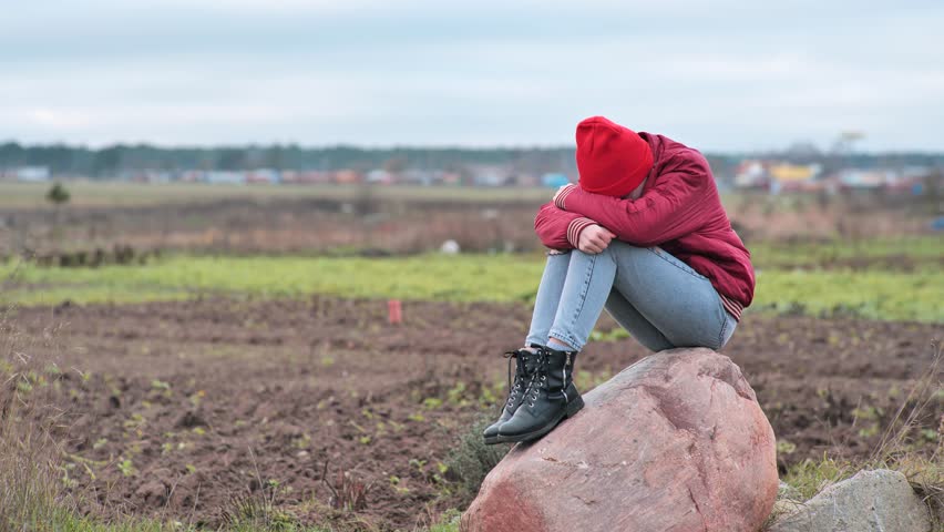 A sad teenage girl sits on a rock in a field and cries.