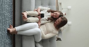 Happy mother hugging daughter kid using smartphone on home couch, talking to child, smiling, enjoying motherhood, family leisure, online communication. Mom and girl sharing mobile phone. Vertical shot
