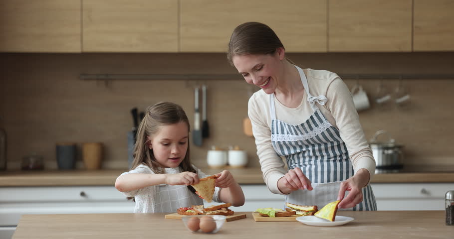 Happy mom and daughter kid wearing aprons, preparing sandwiches for breakfast, cooking in home kitchen together, having fun, laughing. Girl assisting mom, helping to make snacks Royalty-Free Stock Footage #1101194839
