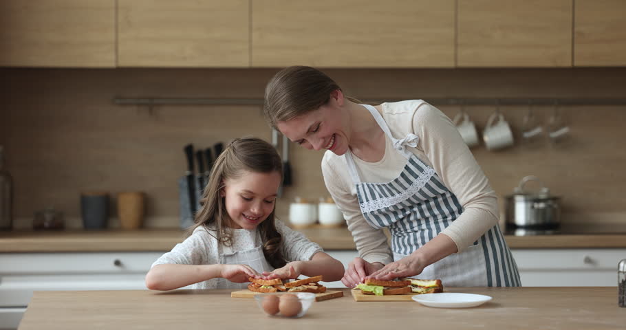 Happy mom and daughter kid wearing aprons, preparing sandwiches for breakfast, cooking in home kitchen together, having fun, laughing. Girl assisting mom, helping to make snacks
