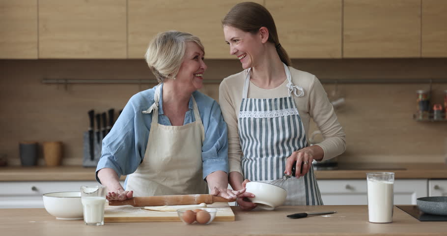 Happy senior mom and adult daughter woman in aprons baking together in kitchen, rolling dough for homemade pastry, bakery food, laughing, enjoying culinary hobby, touching face with floury hand Royalty-Free Stock Footage #1101194847