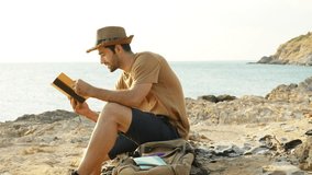 4K Caucasian man traveler reading a book during resting on coastline hill at sunset. Handsome guy enjoy outdoor lifestyle hiking on island mountain on summer holiday vacation. Solo travel concept. - Powered by Shutterstock - Get 15% off with code: PIKWIZARD15