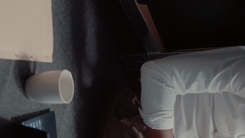 Vertical video: Investigator sitting on floor searching for information in police reports archive, reading witness statements in office. African american agent looking at case files and thinking about