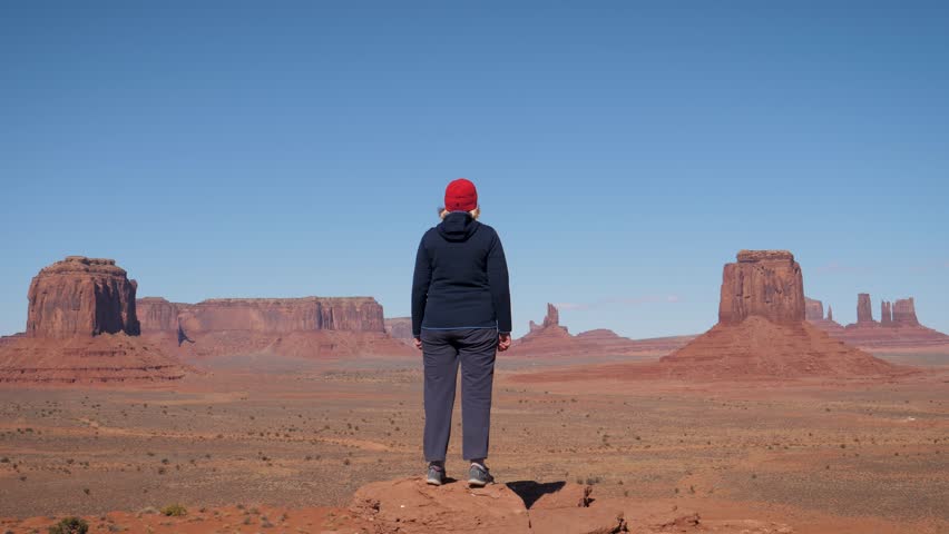 Active woman hiker overweight, standing to edge of cliff at top, raises arms up in air celebrating victory and success. Viewpoint of famous place in monument valley in Western Usa with red rock buttes