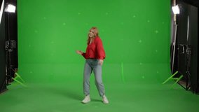 Red-haired white girl listens to music and dances against of green screen. Happy young woman in jeans and a knitted jumper makes a gesture with hands as if swipping the page to the side