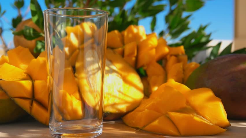Close-up of the fresh mango juice poured in a tall glass next to a mango cut into cubes on a sunny day Royalty-Free Stock Footage #1101207129